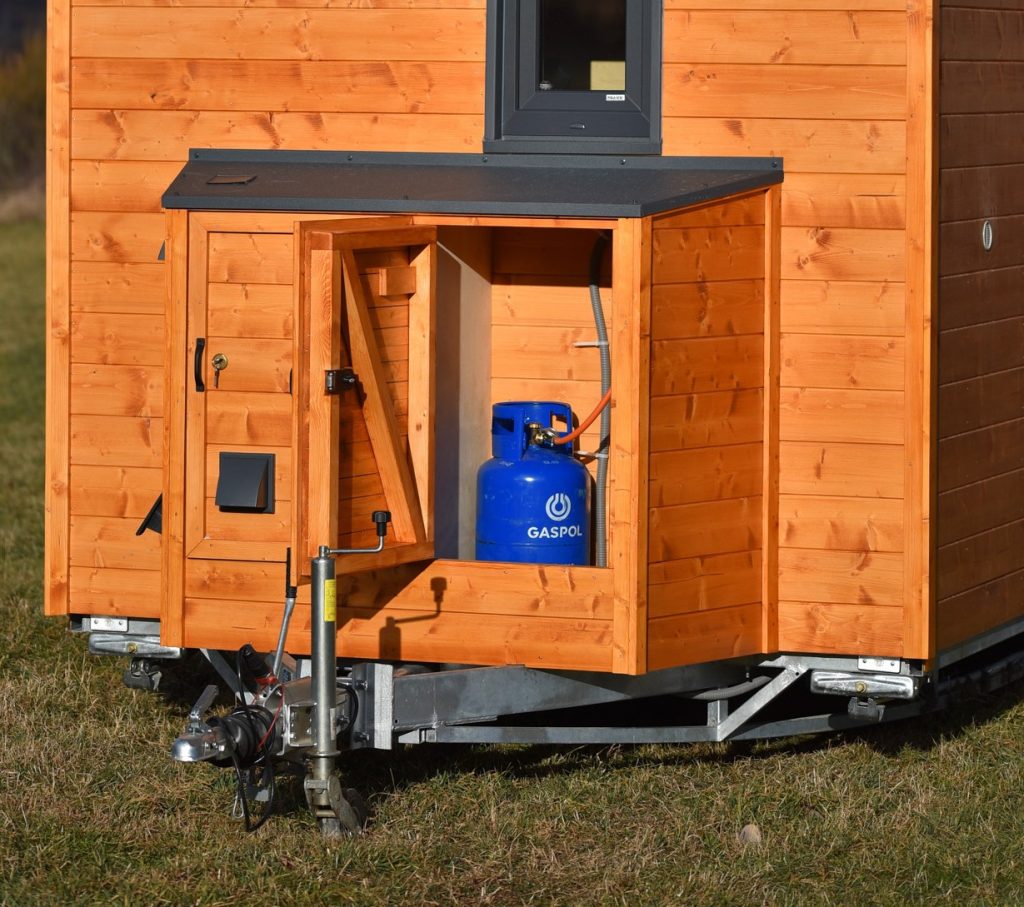Widok ogólny wnętrza tiny house. Na pierwszym planie kuchnia, dalej strefa dzienna ze składanym stolikiem i łóżko na podwyższeniu w głębi. Sufit z oknem dachowym.